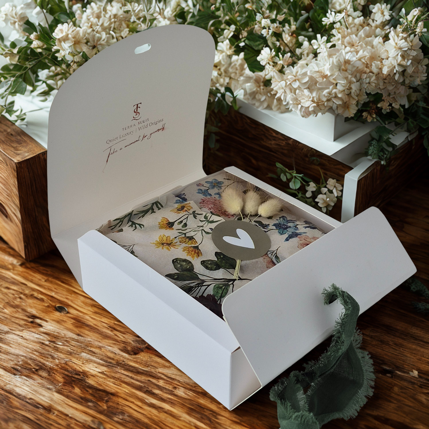 Open white gift box with floral-patterned towel and small items on a wooden surface with flowers in the background.