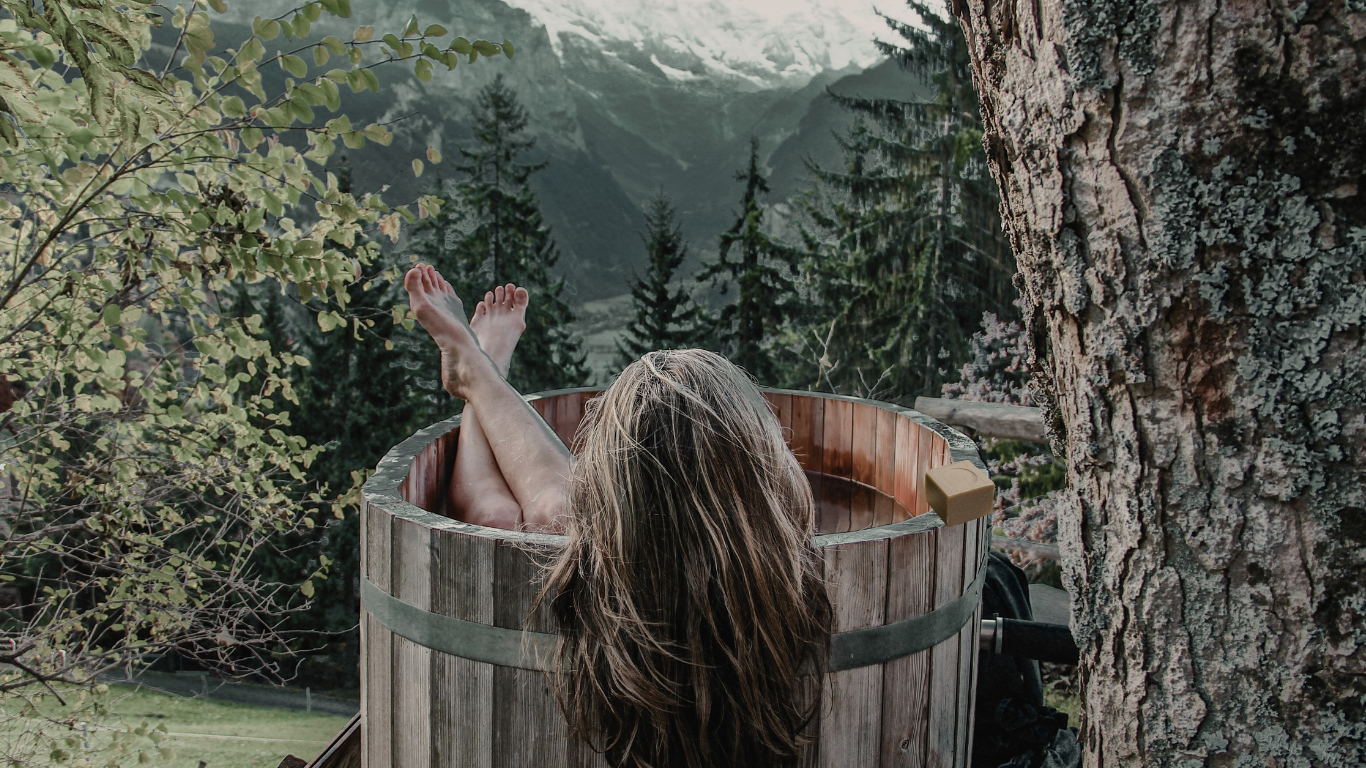 An image of a woman in an outdoor tub looking at the Southern Alps in NZ. A handmade natural soap sits on the side of the tub 